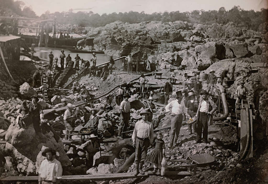 Historic photo of miners at a busy gold mining site with rocky terrain and water. Numerous men and boys, dressed in early 20th-century clothing, are scattered across the scene, working with tools and machinery. Trees are visible in the background.