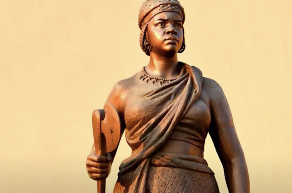 A bronze statue of a woman in traditional attire, holding a paddle or oar. She wears a headdress and a patterned skirt. The background is a plain creamy-yellow wall.