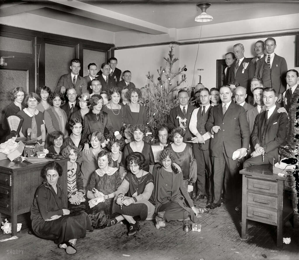 A large group of men and women in 1920s attire posing in an office around a decorated Christmas tree. Some are seated, while others stand, and a few hold drinks. The setting has vintage office furniture and a festive atmosphere.