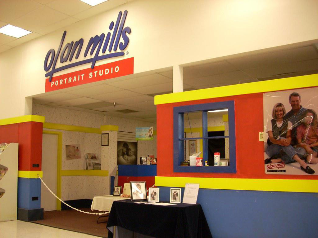 The image shows the entrance to an Olan Mills portrait studio with a colorful facade. A large sign displays "Olan Mills" in blue letters. Photo samples and a family portrait are visible near the entrance.