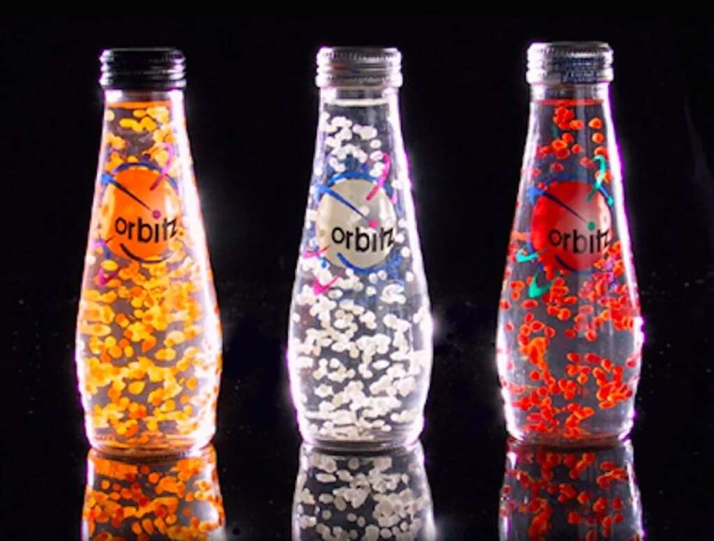 Three bottles of Orbitz drink with colored floating beads are displayed against a black background. The bottles are lit from behind, highlighting the orange, clear, and red liquid contents with their respective colored beads.