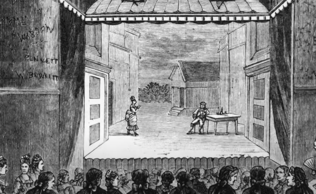 A black and white illustration of a theater stage with two actors. One stands to the left, holding an object, while the other sits at a table to the right. The audience is seated in the foreground, and buildings are visible in the backdrop.