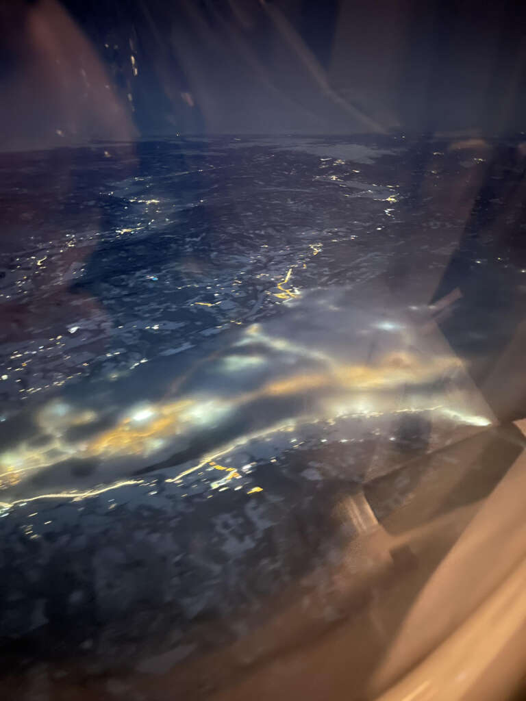 Aerial view of a city at night from an airplane window. The city lights form a network of glowing patterns, with some areas appearing brighter. The reflection on the window adds a soft blur to the overall scene.