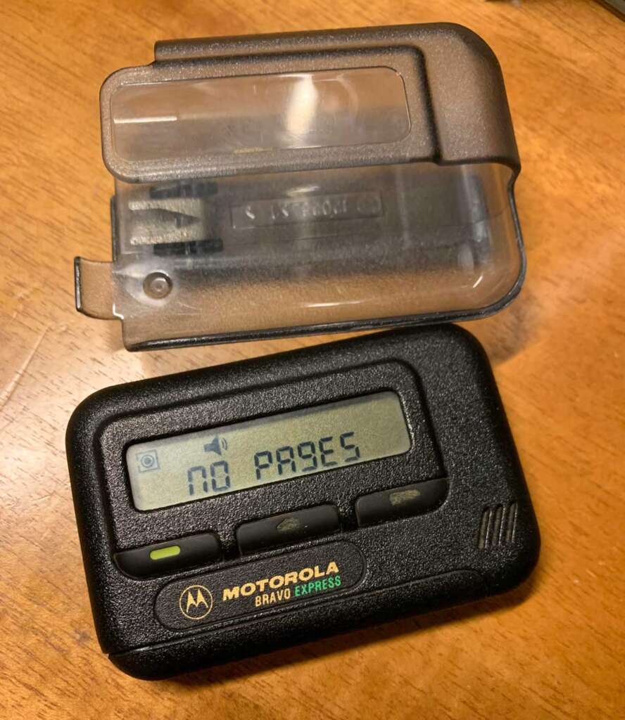 A Motorola Bravo Express pager with a "NO PAGES" message displayed on its screen. It is placed on a wooden surface alongside its transparent plastic cover.