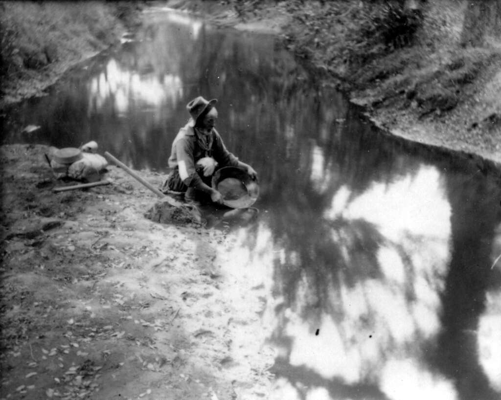 A person wearing a hat is crouching by a river with a pan, likely panning for gold. There are some bags and tools beside them. The water reflects trees from the surrounding landscape. The scene is in black and white.