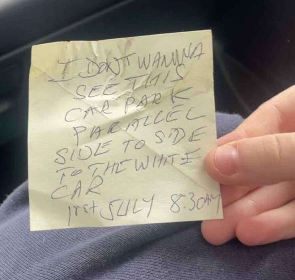 A crumpled yellow sticky note with handwritten text reads: "I don't wanna see this car park parallel side to side to the white car. 1st July 8:30 AM." The note is held by a person wearing a blue garment.