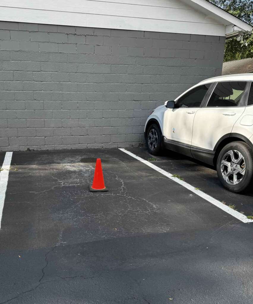 A white SUV is parked in the last spot of a parking lot, next to a gray wall. An orange traffic cone is placed in the adjacent empty parking space. The ground is asphalt, and there are painted white lines marking the parking spaces.