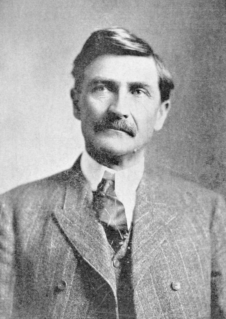 Black-and-white portrait of a man with short hair and a mustache, wearing a three-piece suit with a tie. The backdrop is plain, highlighting his formal attire and composed expression.