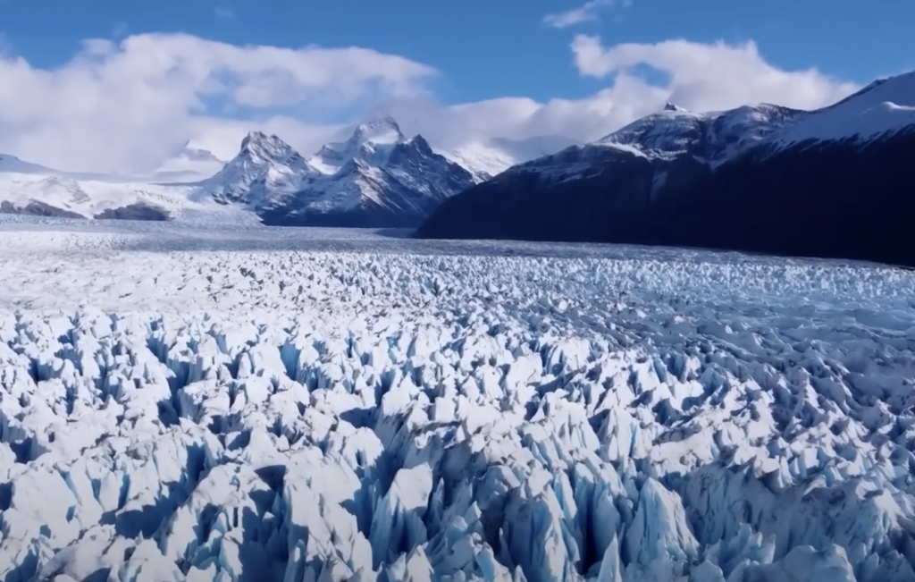 A vast expanse of jagged, blue-tinged ice formations stretches across a glacier beneath a bright blue sky. Snow-capped mountains rise in the background, partially covered by soft, white clouds.