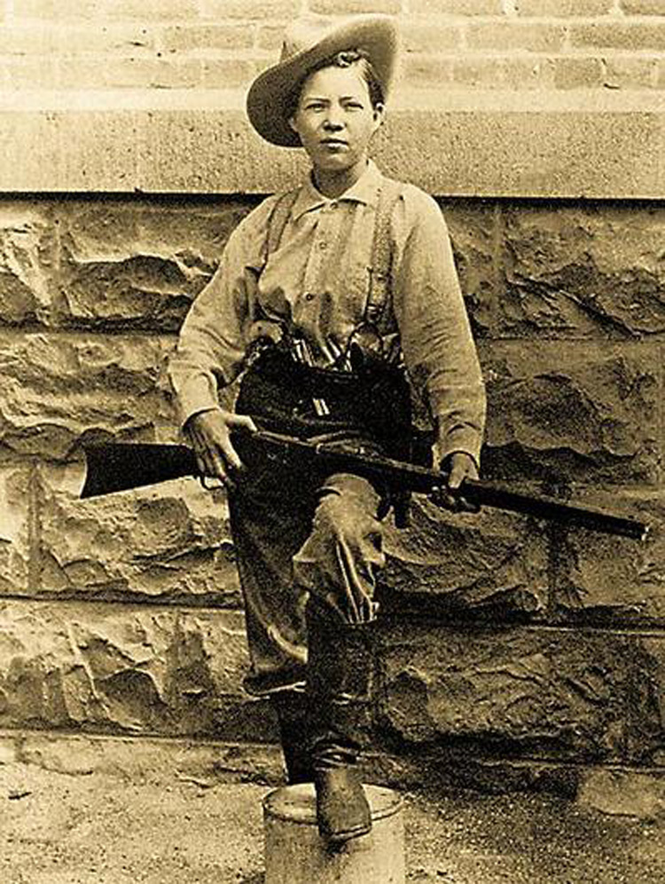 A historical black and white photo of a person wearing a wide-brimmed hat, long-sleeve shirt, belt, and boots, standing confidently with one foot on a low column, holding a rifle. They are posed against a stone wall background.