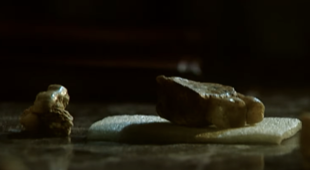 Close-up of two nuggets placed on a dark surface. One smaller nugget is positioned to the left, while a larger, irregularly-shaped nugget rests on a piece of light-colored material to the right. Shadows and light create a dramatic effect.