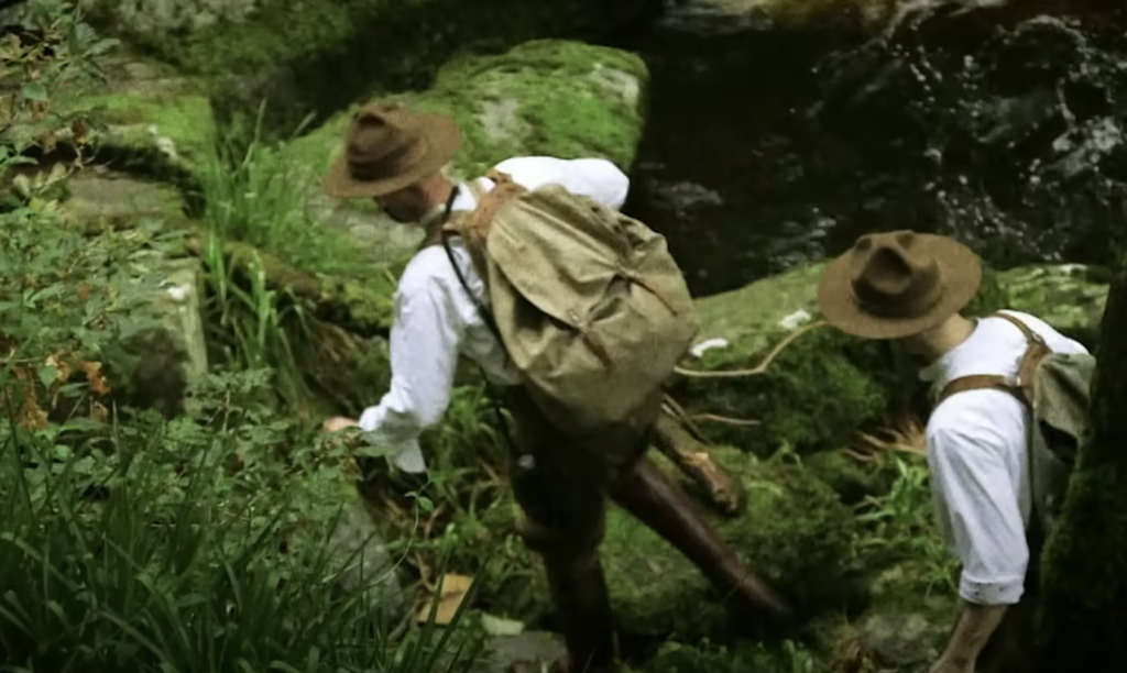 Two people in vintage attire, wearing hats and suspenders, navigate a moss-covered rocky terrain near a stream. They appear to be exploring a forested area.