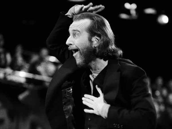 A black and white image of a man with long hair and a beard, animatedly gesturing with one hand on his head and the other on his chest. He appears to be speaking or performing on stage in front of an audience.