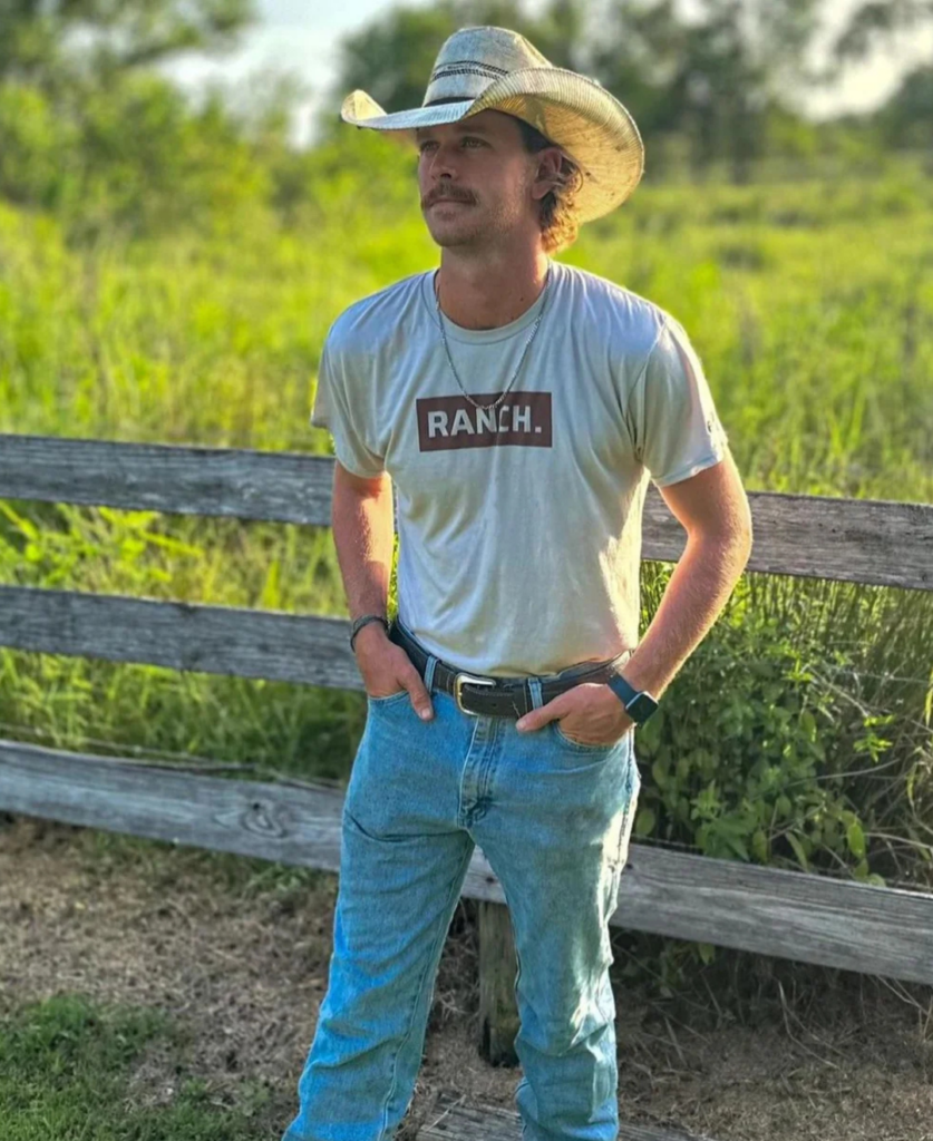 A man wearing a cowboy hat stands outdoors by a wooden fence. He is dressed in a "RANCH" T-shirt, jeans, and a watch, with hands in his pockets. The background features greenery and a sunny sky.