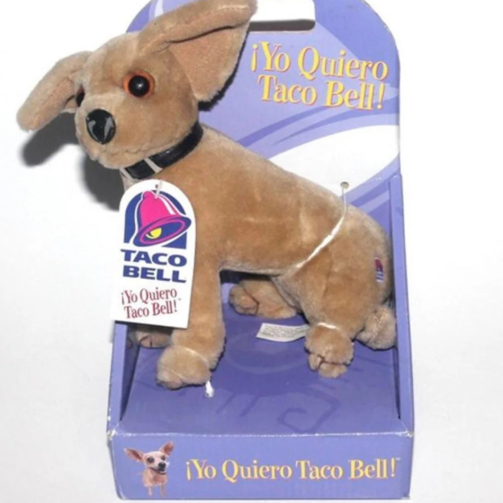 A plush chihuahua toy with large ears and a black collar sits in purple packaging featuring Taco Bell branding and the text "¡Yo Quiero Taco Bell!" A tag is attached to the collar with the same text and logo.