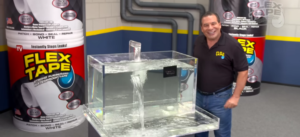 Man in a black polo shirt stands next to a clear tank with water spilling from a large rectangular hole in the side. The hole is sealed with clear tape. Large Flex Tape rolls are visible in the background.