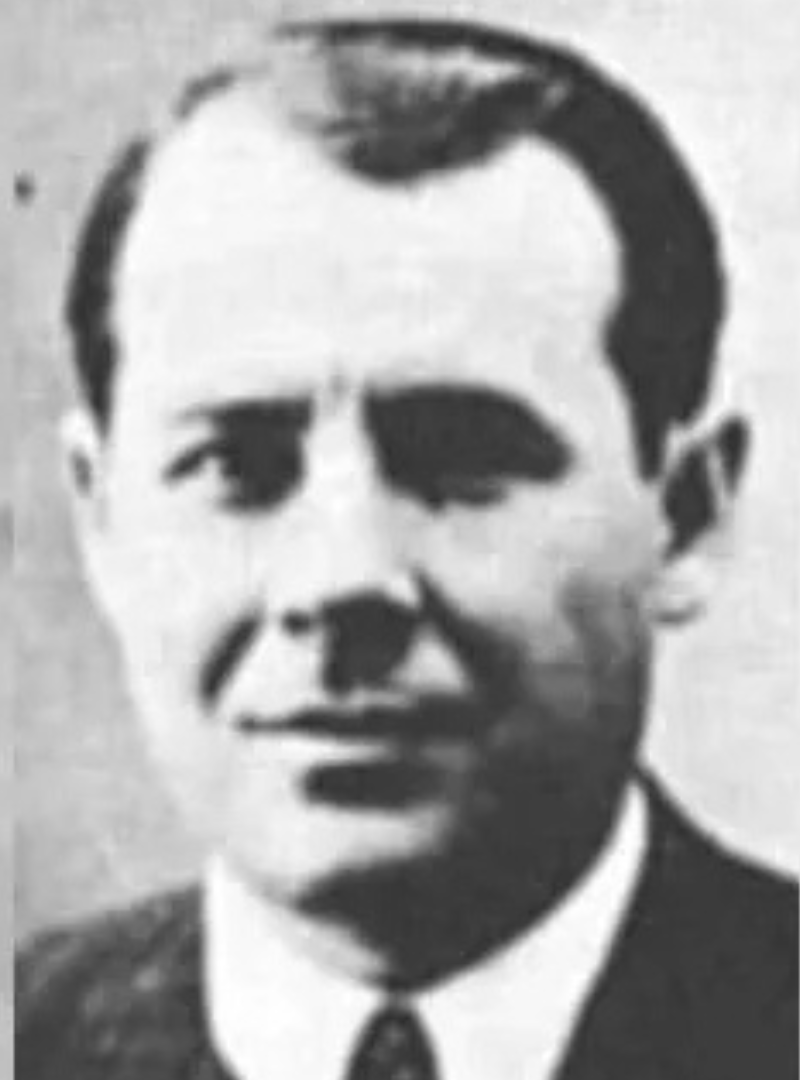 Black and white photo of a man wearing a suit and tie. He has short, slicked-back hair and an unreadable expression. The background is plain and the image has a portrait format.