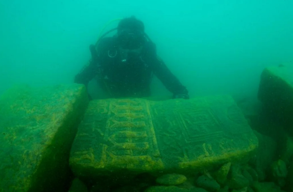 A scuba diver examines ancient stone tablets underwater. The tablets are engraved with intricate patterns and inscriptions, partially covered in algae. The surroundings have a greenish tint from the water.