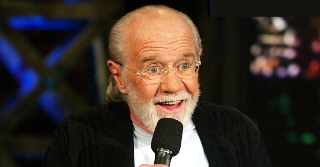 An older man with glasses and a beard holds a microphone while speaking animatedly. He is wearing a black jacket over a white shirt, and the background is blurred with colorful lights.