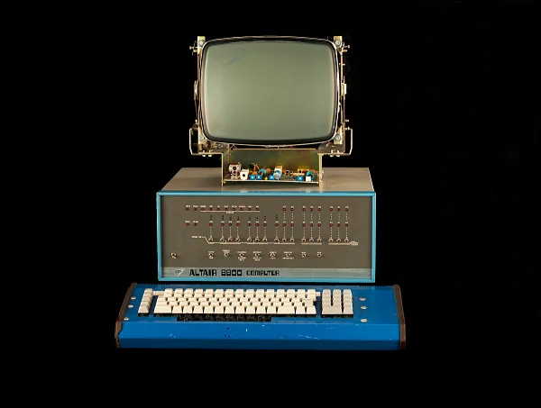 A vintage Altair 8800 computer setup, featuring a green monochrome monitor, a large mainframe with switches and lights, and a blue keyboard with a numeric keypad, is displayed against a black background.