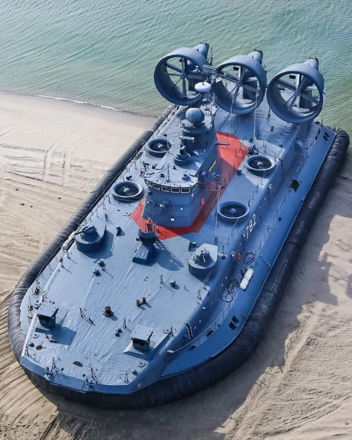 An aerial view of a large military hovercraft with three propellers, resting on a sandy beach near the water. Its gray and blue structure features various hatches and markings.