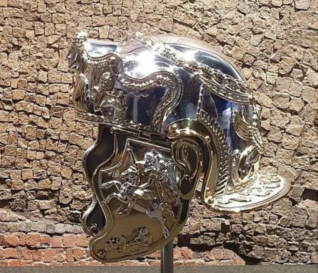 A shiny, ornate Roman cavalry helmet with intricate silver and gold detailing, featuring a relief of a horse and rider on the cheek guard. It's displayed against a textured stone wall.