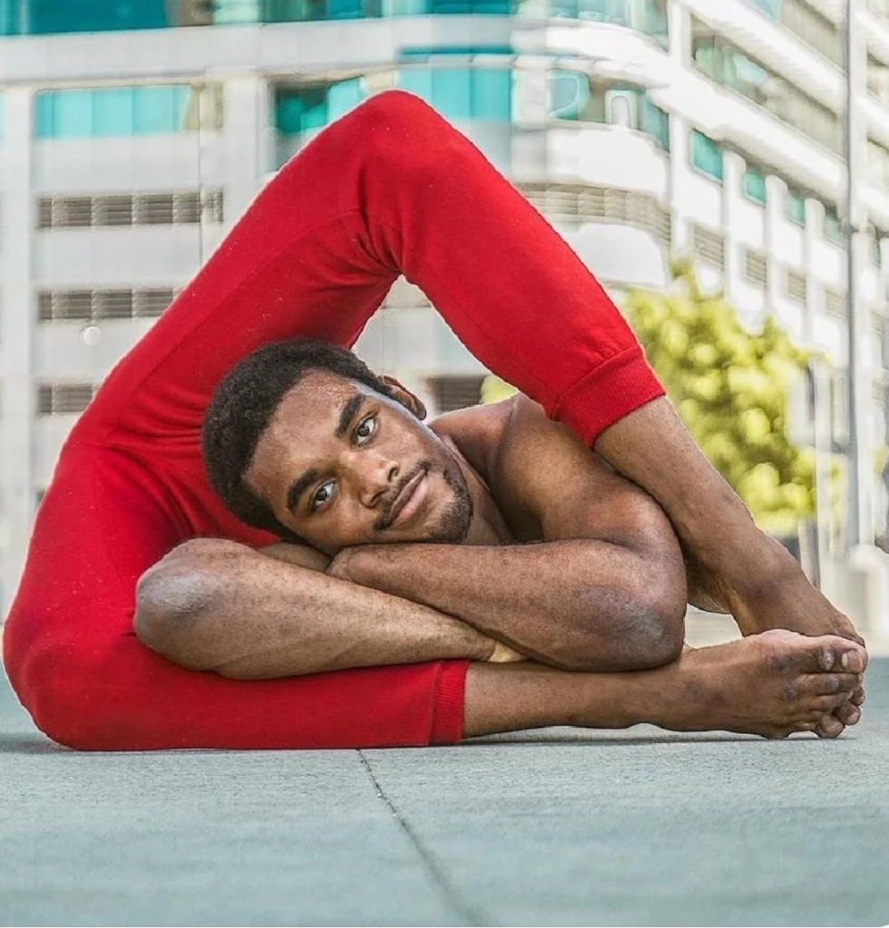 A person wearing red pants is performing an impressive contortion pose, reclining with their legs bent over their shoulders. An urban outdoor setting with buildings in the background can be seen.