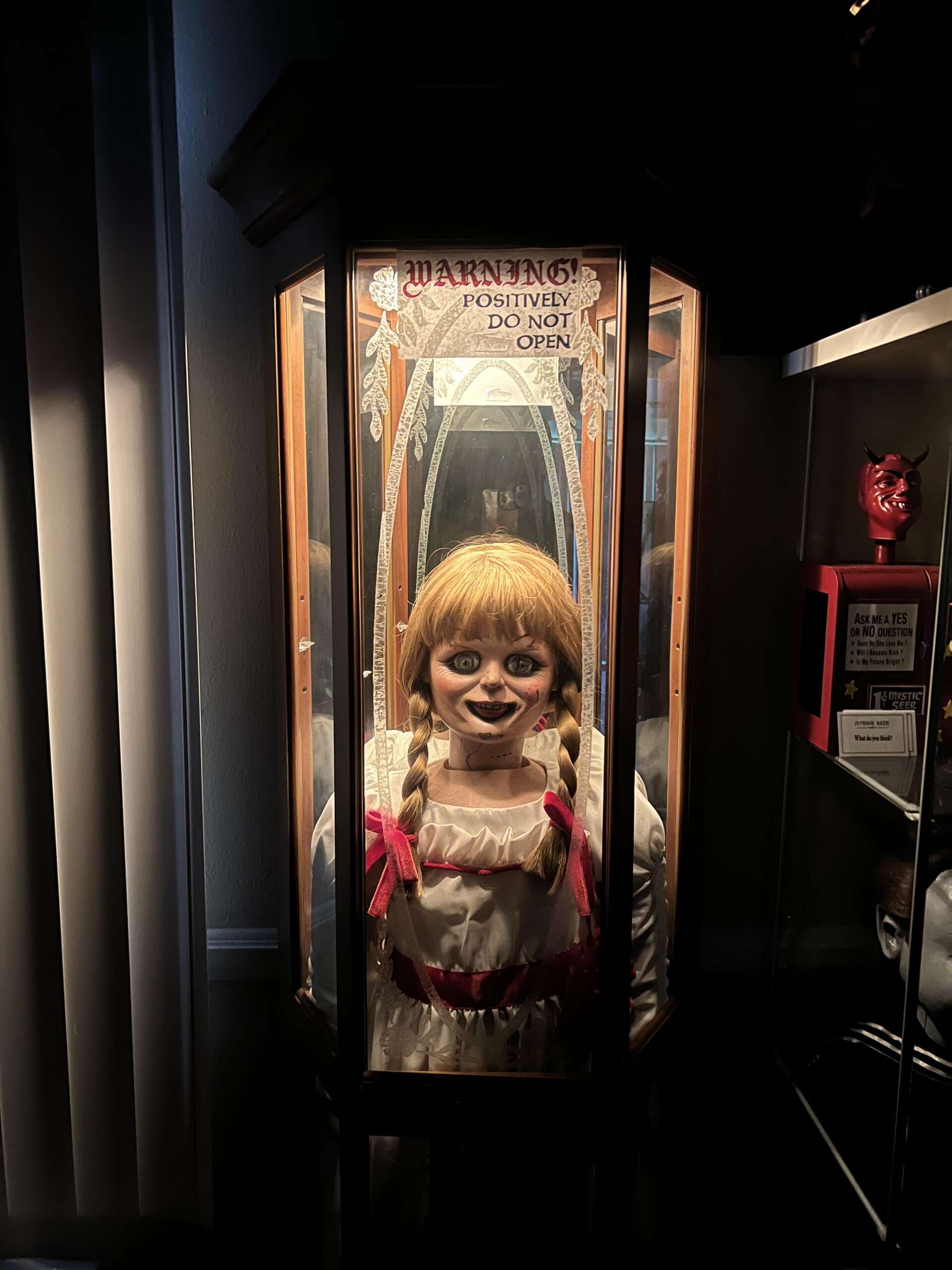 A doll with red braids and a white dress is enclosed in a wooden and glass case. A sign above reads, "Warning: Positively Do Not Open." The setting is dark, with a red devil mask displayed nearby.