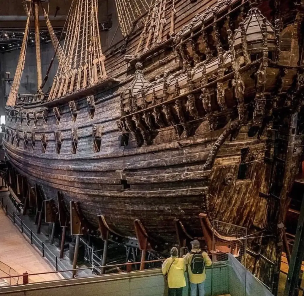 A large, well-preserved wooden ship with intricate carvings is displayed in a museum. Three people stand in the foreground, observing the ship's impressive size and details.