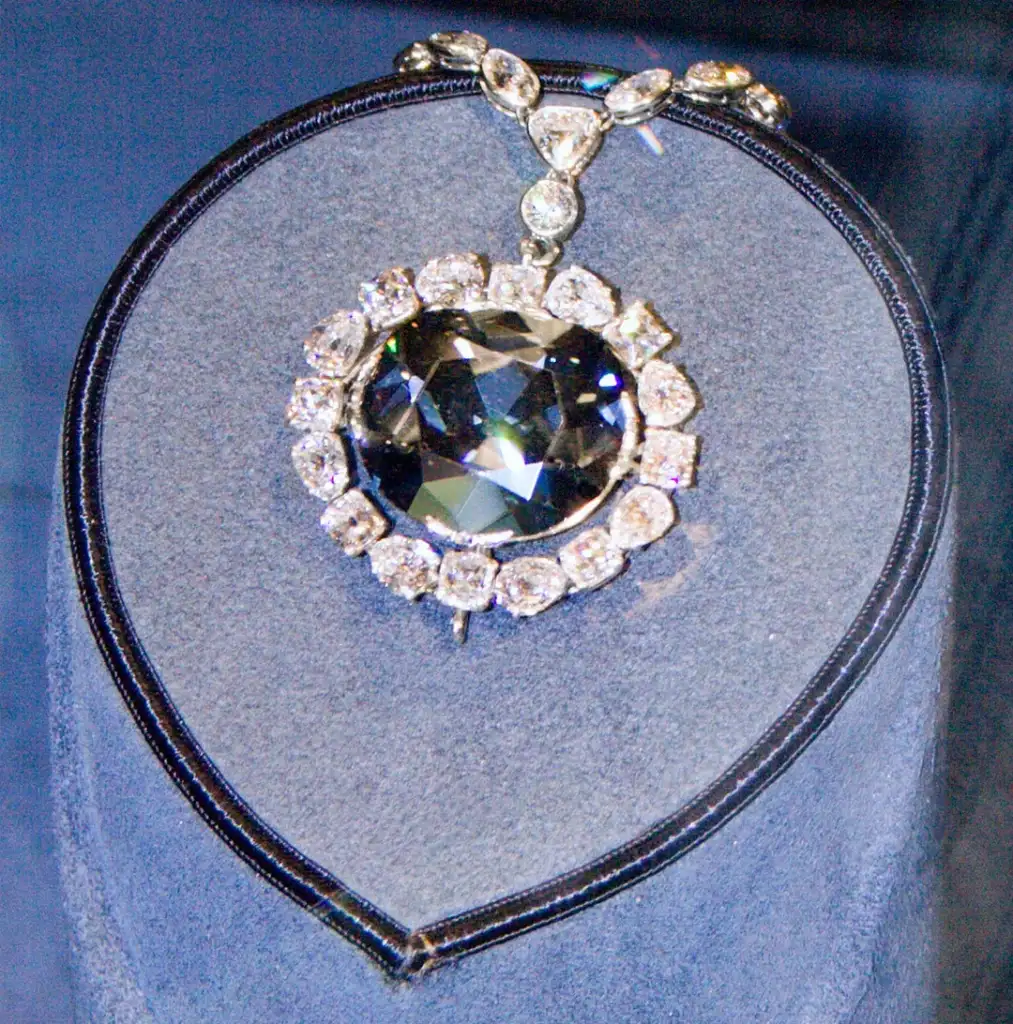 A large, dark blue diamond pendant set in a necklace with a ring of clear, sparkling diamonds. The necklace is displayed on a dark blue velvet stand.