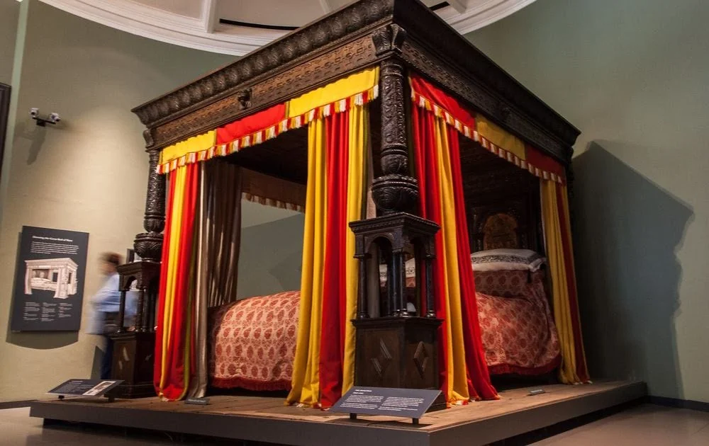 A large, ornately carved four-poster bed with rich red and yellow drapes is displayed in a museum setting. The bed features intricate woodwork and stands on a raised platform. A person is visible in the background.