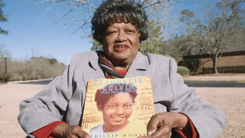 Elderly woman sitting outdoors, smiling, and holding a book titled "Claudette Colvin: Twice Toward Justice" featuring a young girl's photo on the cover. Trees and a building in the background.