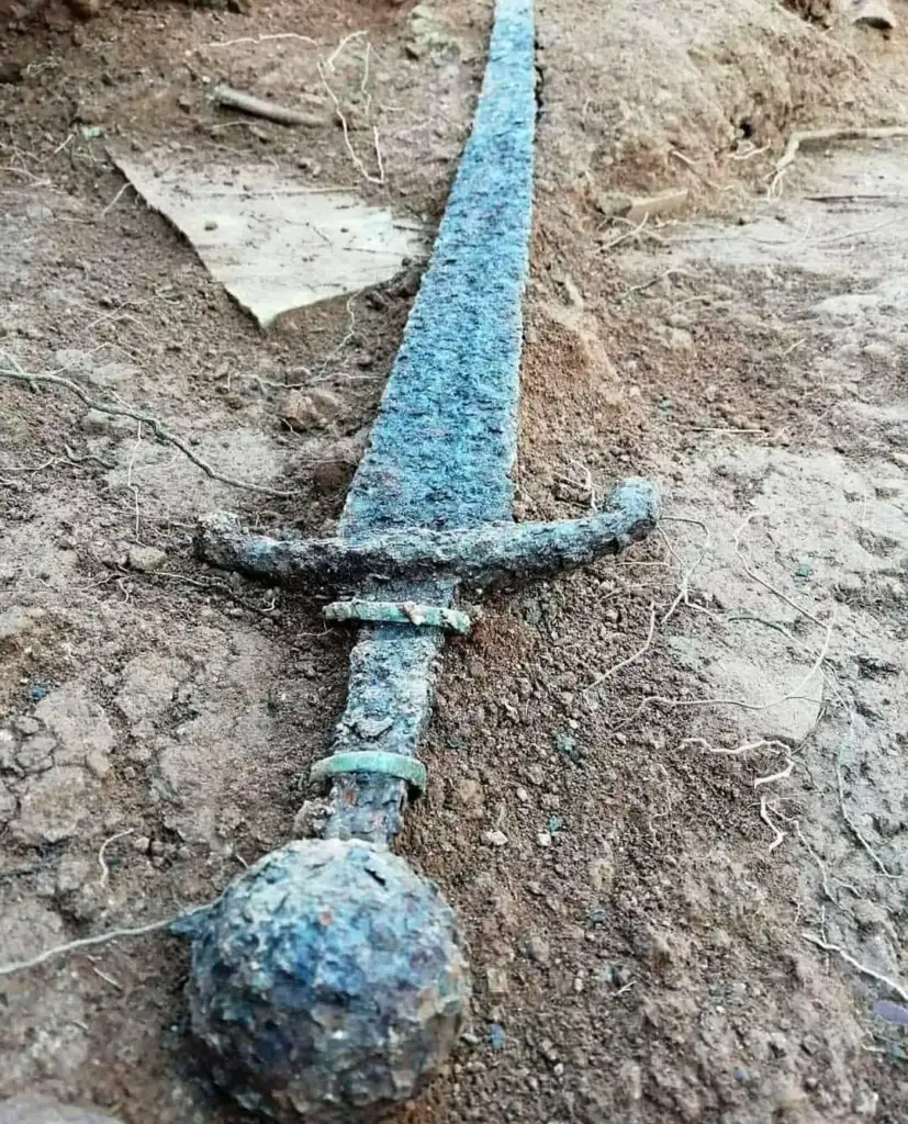 A rusted, ancient sword partially buried in dirt. The sword features a noticeable hilt and a long, narrow blade, showing signs of corrosion and age.