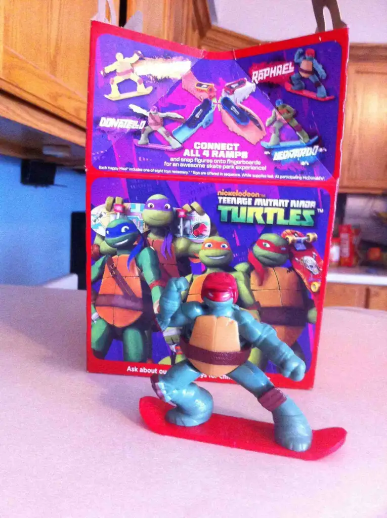 An action figure of a Teenage Mutant Ninja Turtle stands on a red skateboard in front of a colorful cardboard backdrop. The backdrop features illustrations of the four turtles: Donatello, Michelangelo, Raphael, and Leonardo.