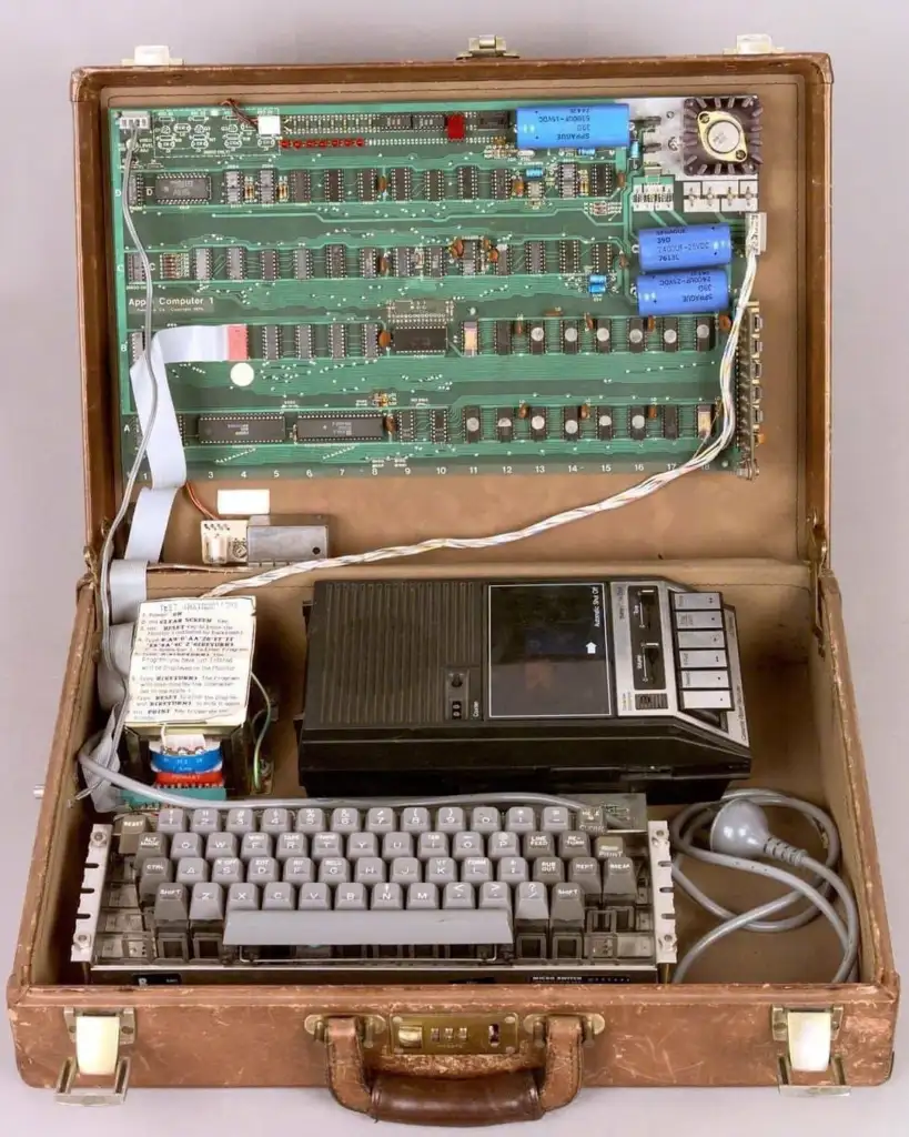 Open briefcase displaying vintage computer hardware, featuring a large green circuit board with various components, a tape recorder, keyboard, and connected cables. The retro setup is housed in a brown leather case with a handle.