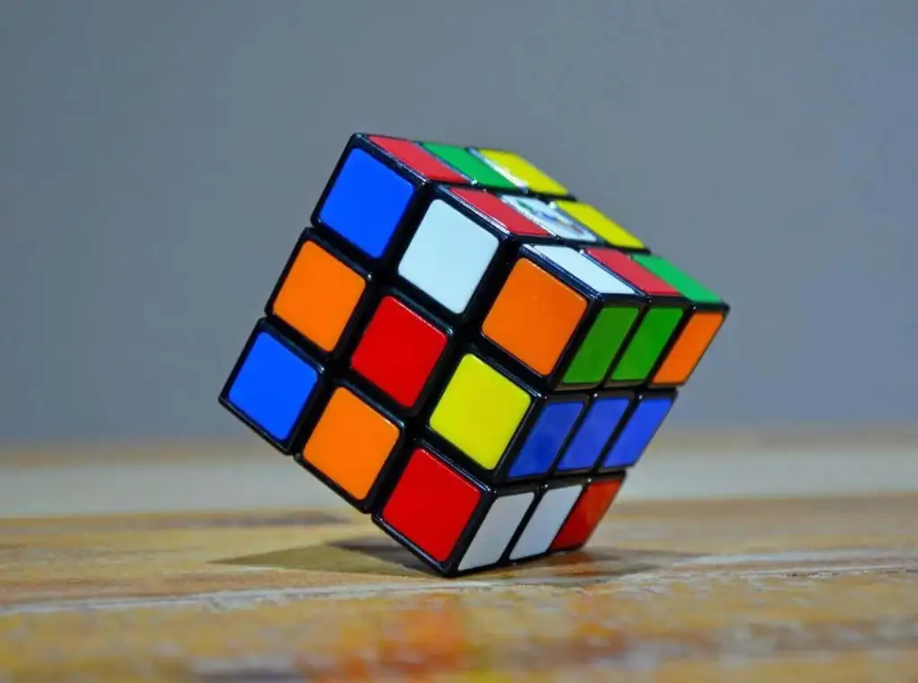 A partially solved Rubik's Cube resting on a wooden surface with a gray background. The cube features mixed colors on its visible sides, including blue, red, yellow, orange, green, and white.