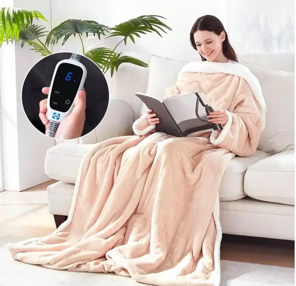 A woman sits on a couch wrapped in a beige heated blanket with white trim, reading a book. A circular inset shows a close-up of the blanket’s control device displaying the number six. Green plants are in the background.