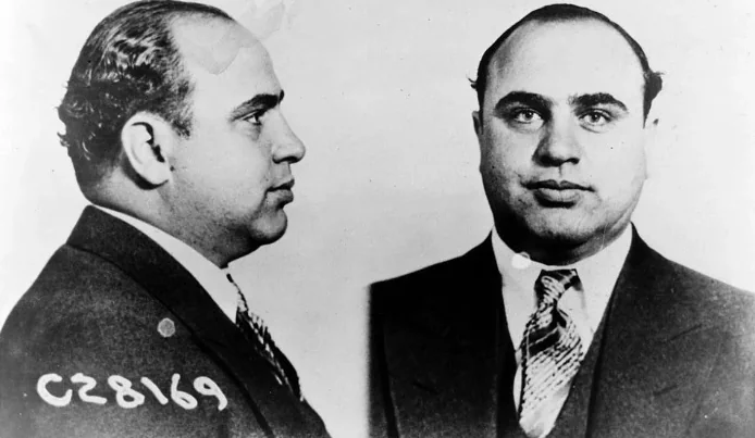 A black-and-white mugshot of a man wearing a suit and tie, shown from a front and side profile. The number "C728169" is visible on the side profile image.