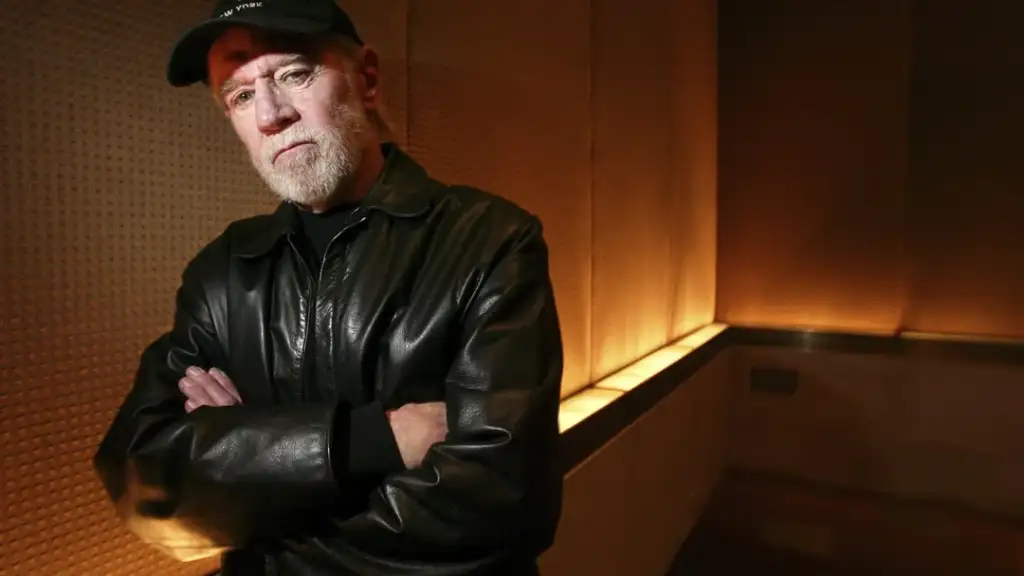 A man with a beard wearing a black leather jacket and cap stands with arms crossed in a dimly lit room with textured walls. The lighting casts a warm glow, creating a contrasting background.