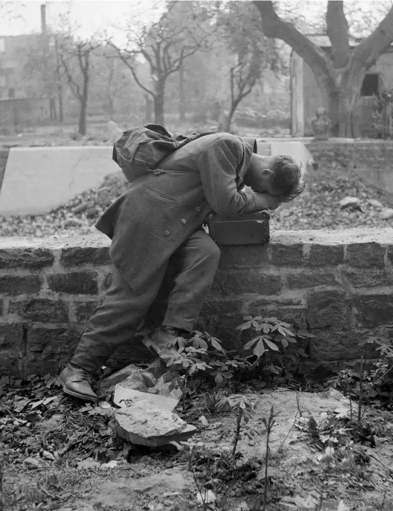 A person in a coat leans over a stone wall, face buried in their arms, with a backpack on their back. The ground is littered with papers, and the background shows trees and blurred buildings. The scene appears somber and contemplative.