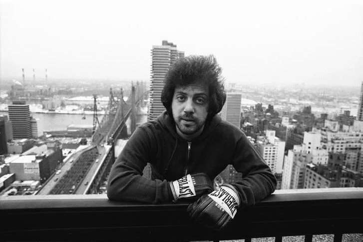 A person in a dark zip-up jacket and boxing gloves leans on a railing overlooking a cityscape. In the background, a bridge stretches across the scene, and tall buildings are visible under a cloudy sky.