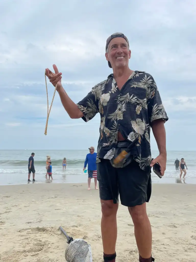 A man in a floral shirt and cap holds a necklace while standing on a beach. He smiles, surrounded by people in the background near the ocean. A metal detector lies on the sand beside him. The sky is overcast.