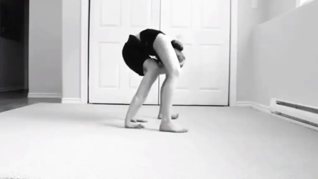 A person performing a contortionist pose in a room with carpeted floor. They are bent forward, legs straight, with their head and shoulders low to the ground, arms wrapped around their legs. The background shows a white door and walls.