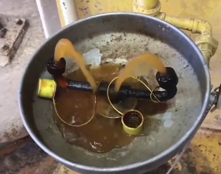 Rusty water flows from two connected pipes into a large metal basin, creating a brown, murky liquid. The setup appears industrial, with visible corrosion and yellow paint on some surfaces.