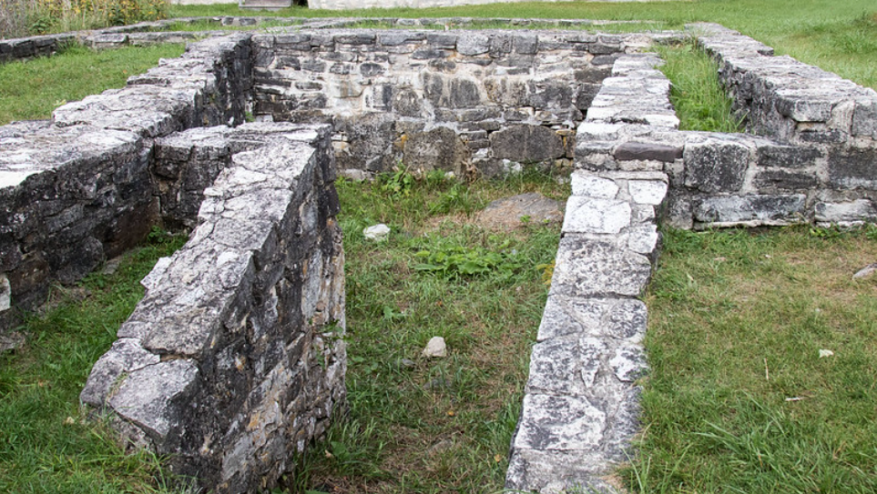 Stone ruins of a small structure with rectangular foundations and low walls, surrounded by grass. The rough stone walls are partially intact, and greenery is growing within the enclosure, suggesting an old, abandoned site.