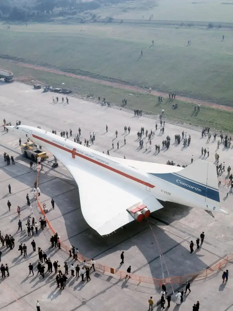 Aerial view of a Concorde aircraft on display at an airfield, surrounded by a crowd of people. The area is bordered by orange barriers, and there are grassy fields in the background. The airplane has a distinctive delta wing design.