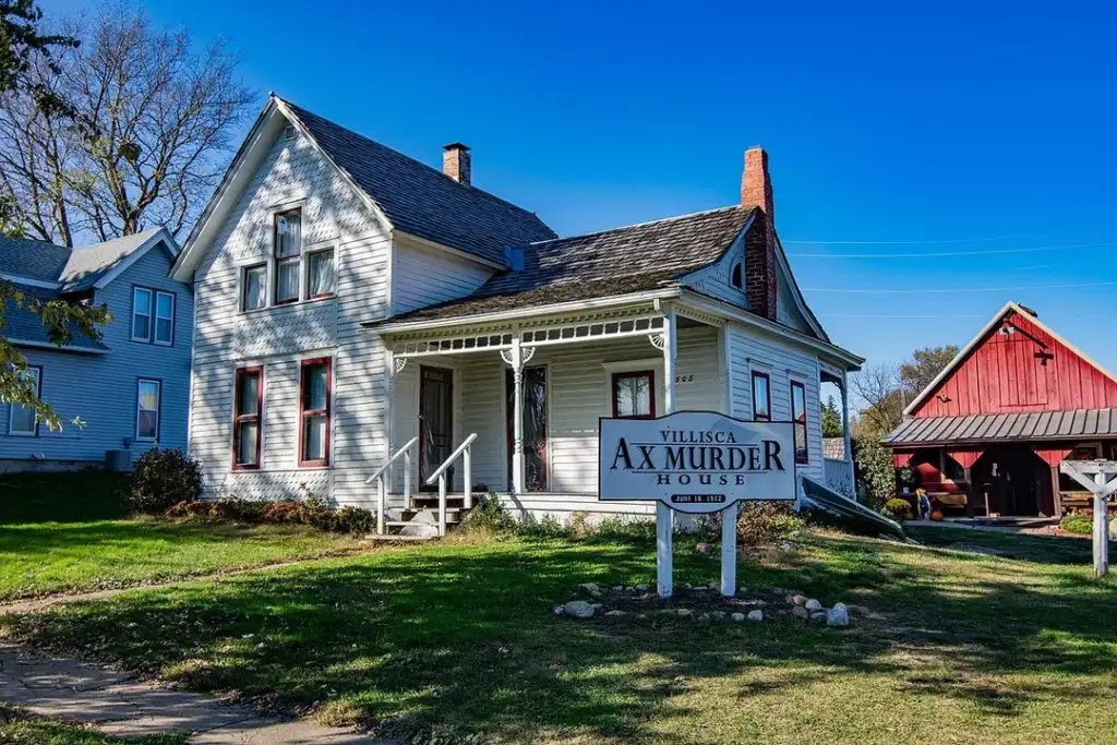 White two-story house with dark shingles and a covered porch, labeled "Villisca Ax Murder House" on a white sign in the front yard. A red barn is visible in the background under a clear blue sky.