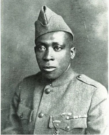 A black and white portrait of a soldier wearing a uniform and a military cap. He has a serious expression and visible medals on his uniform. The background is plain.