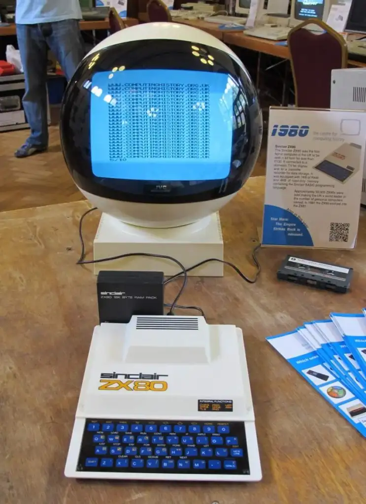 A retro computer setup featuring a Sinclair ZX80 with a white case and blue keypad, connected to a futuristic, round monitor displaying a blue screen with a pattern. Nearby, informational pamphlets are placed on the wooden table.