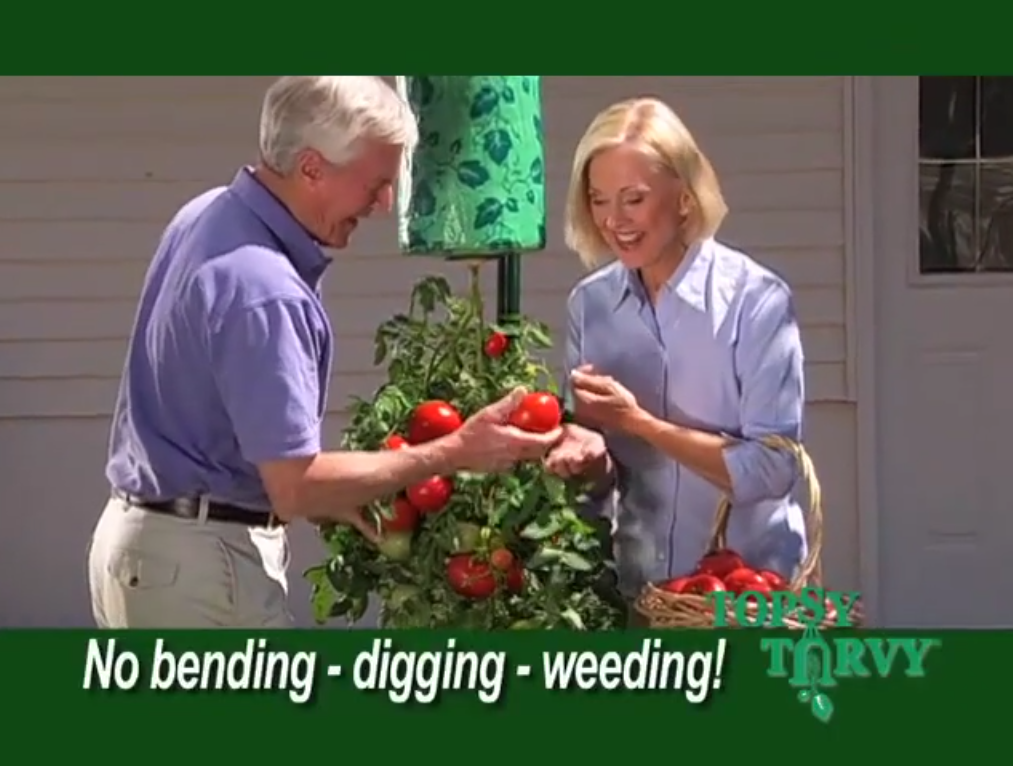 An elderly couple happily picks ripe tomatoes from an upside-down planter outside a house. They are wearing casual clothes, and the text on the image reads "No bending - digging - weeding! Topsy Turvy.